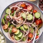 A vibrant Mediterranean chopped salad in a bowl showcases sliced cucumbers, cherry tomatoes, red onion rings, kalamata olives, and feta cheese crumbles. A serving spoon rests inside. Surrounding bowls offer extra cucumber slices, olives, and feta cheese for garnish or indulgence.