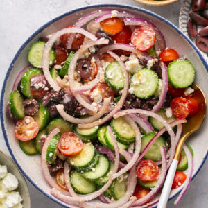 A vibrant Mediterranean chopped salad in a bowl showcases sliced cucumbers, cherry tomatoes, red onion rings, kalamata olives, and feta cheese crumbles. A serving spoon rests inside. Surrounding bowls offer extra cucumber slices, olives, and feta cheese for garnish or indulgence.