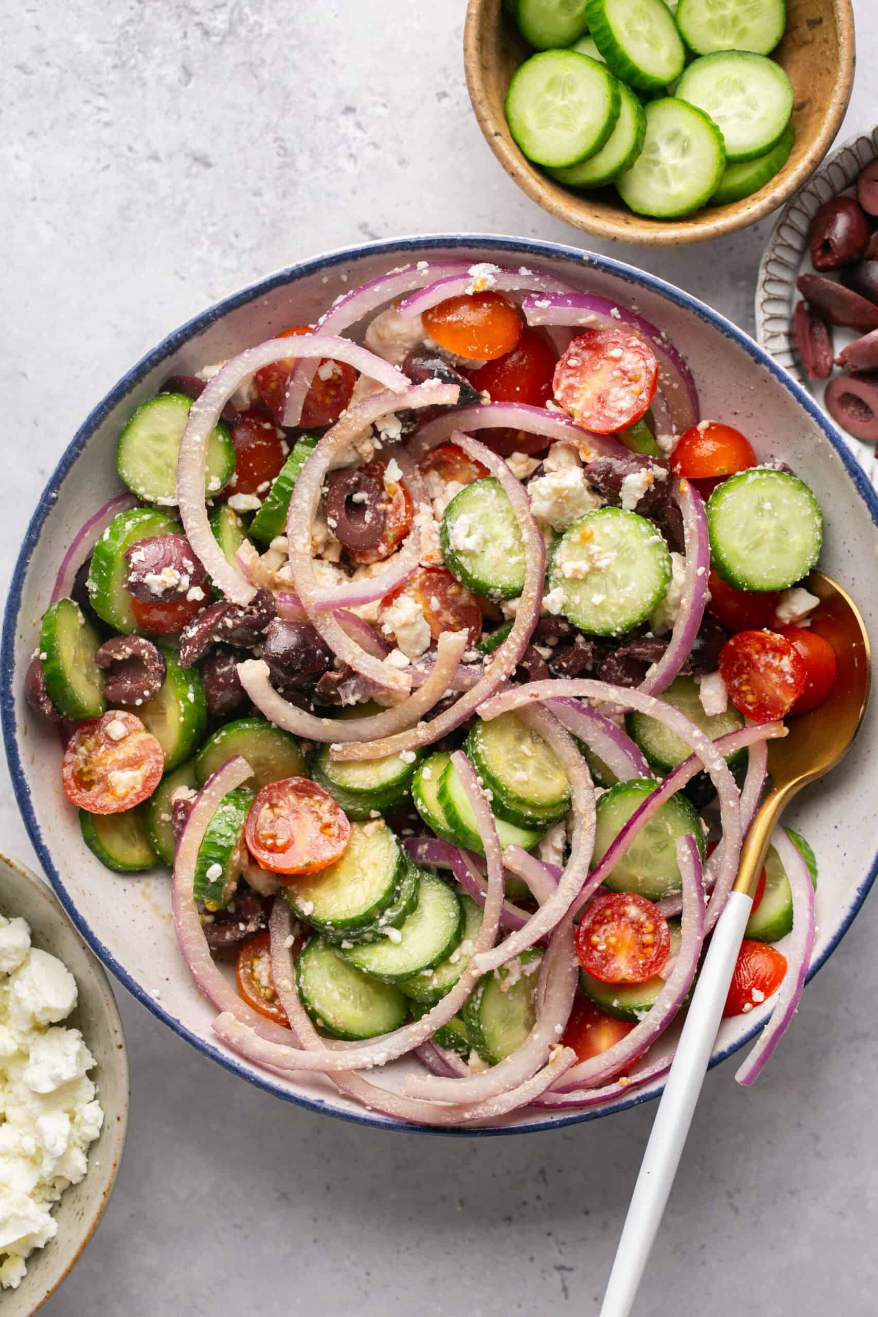 A vibrant Mediterranean chopped salad in a bowl showcases sliced cucumbers, cherry tomatoes, red onion rings, kalamata olives, and feta cheese crumbles. A serving spoon rests inside. Surrounding bowls offer extra cucumber slices, olives, and feta cheese for garnish or indulgence.