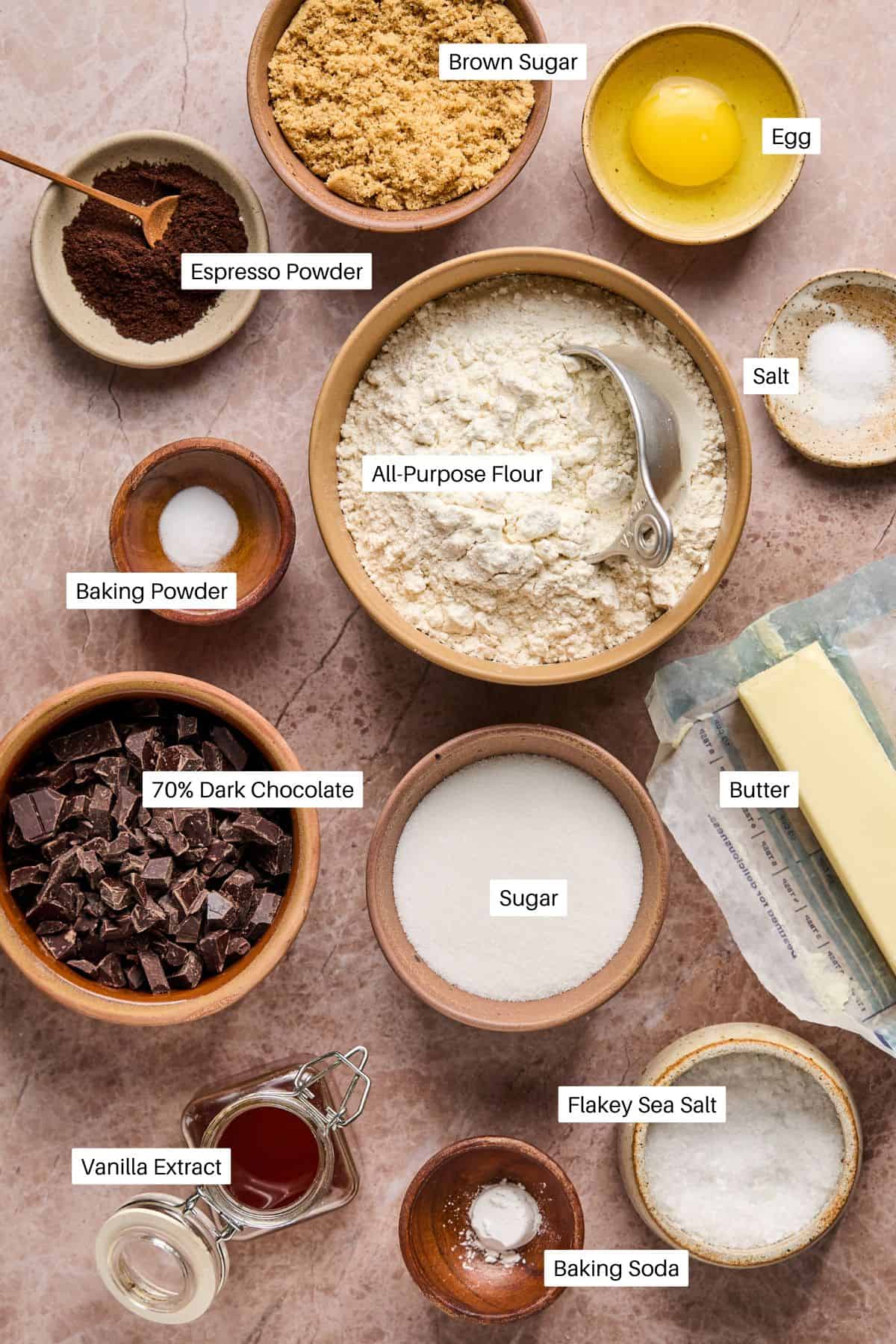 A flat lay of coffee cookie ingredients, featuring bowls of flour, sugar, brown sugar, chocolate chunks, espresso powder, baking powder, baking soda, salt, and eggs. Accompanied by butter and a bottle of vanilla extract.