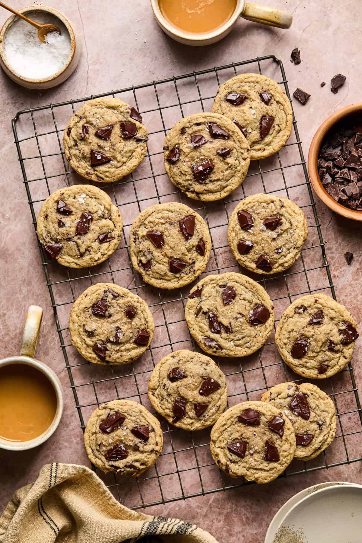 A cooling rack holds 13 chocolate chip cookies. Surrounding the rack are bowls of chocolate chunks, salt, and two mugs of coffee. A beige cloth is partially visible at the bottom, all set on a pinkish stone surface.