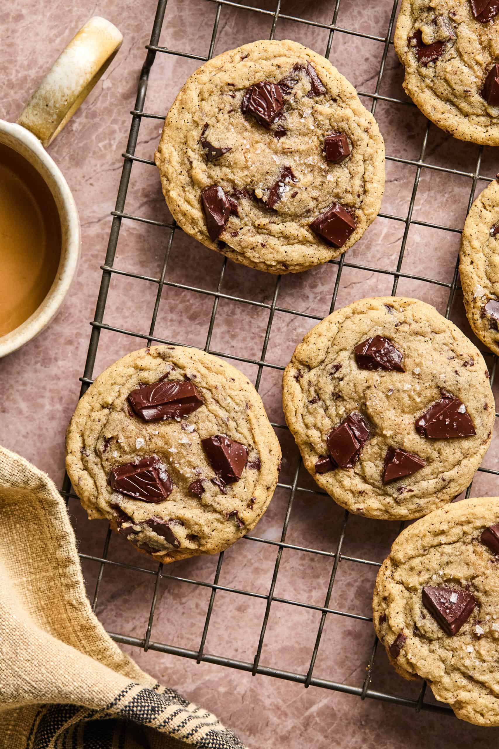 Chocolate chip cookies cool on a wire rack next to a steaming mug of richly brewed coffee. A cozy cloth napkin is placed nearby, creating a warm and inviting scene that perfectly pairs coffee with cookies.