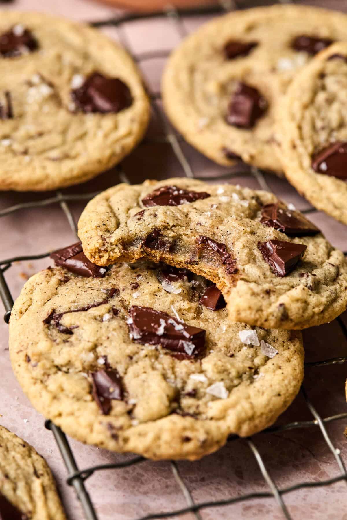 Close-up of chocolate chip coffee cookies on a wire rack. One cookie is stacked on another with a bite taken out, revealing a soft interior with chunks of chocolate. Sprinkles of sea salt are visible on top, adding texture and flavor.