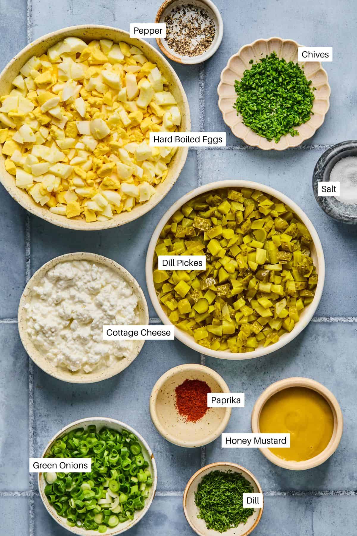 A top-down view features an array of ingredients in bowls on a gray surface, perfect for creating a Cottage Cheese Egg Salad. Labeled are hard-boiled eggs, dill pickles, cottage cheese, honey mustard, with smaller bowls of chives, green onions, paprika, dill, pepper, and salt.