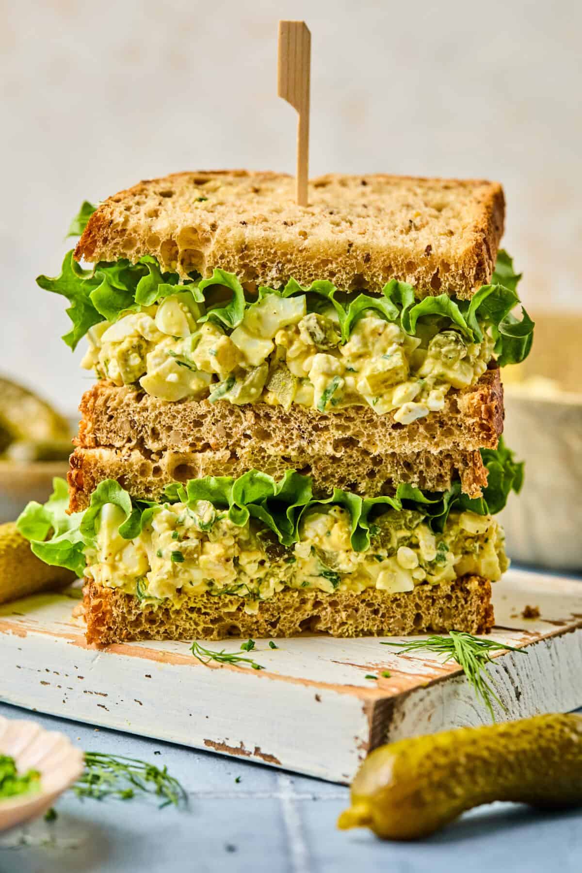 A stack of chicken and cottage cheese egg salad sandwich halves on whole wheat bread with crisp green lettuce. A wooden skewer holds them together. They're artfully arranged on a white rustic board, surrounded by pickles and fresh dill sprigs.