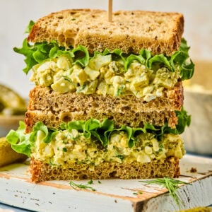 A stack of chicken and cottage cheese egg salad sandwich halves on whole wheat bread with crisp green lettuce. A wooden skewer holds them together. They're artfully arranged on a white rustic board, surrounded by pickles and fresh dill sprigs.