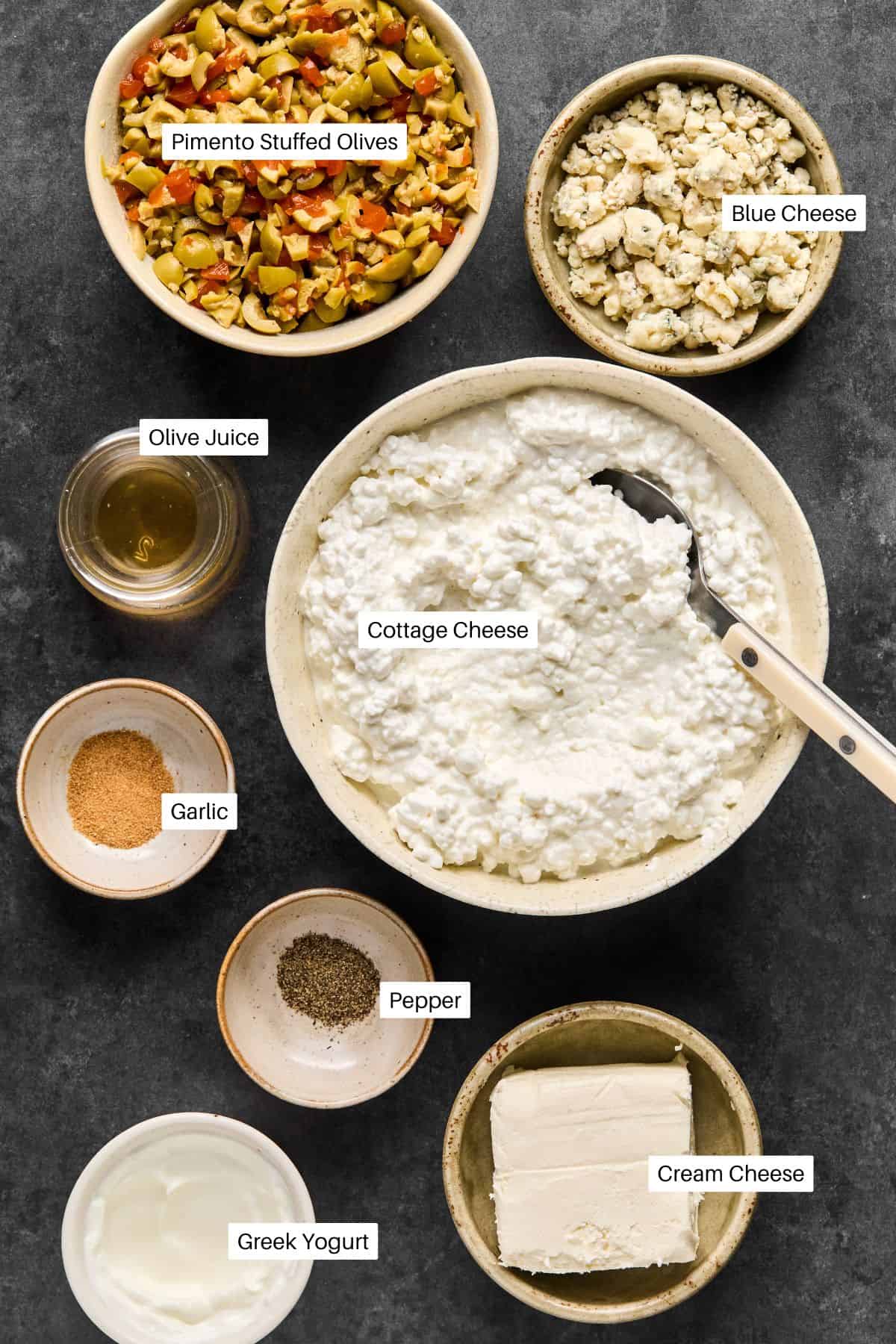 A dark surface showcases bowls of various ingredients for a dirty martini dip recipe: pimento stuffed olives, blue cheese, cottage cheese, olive juice, garlic, pepper, Greek yogurt, and cream cheese. A spoon rests in the cottage cheese.