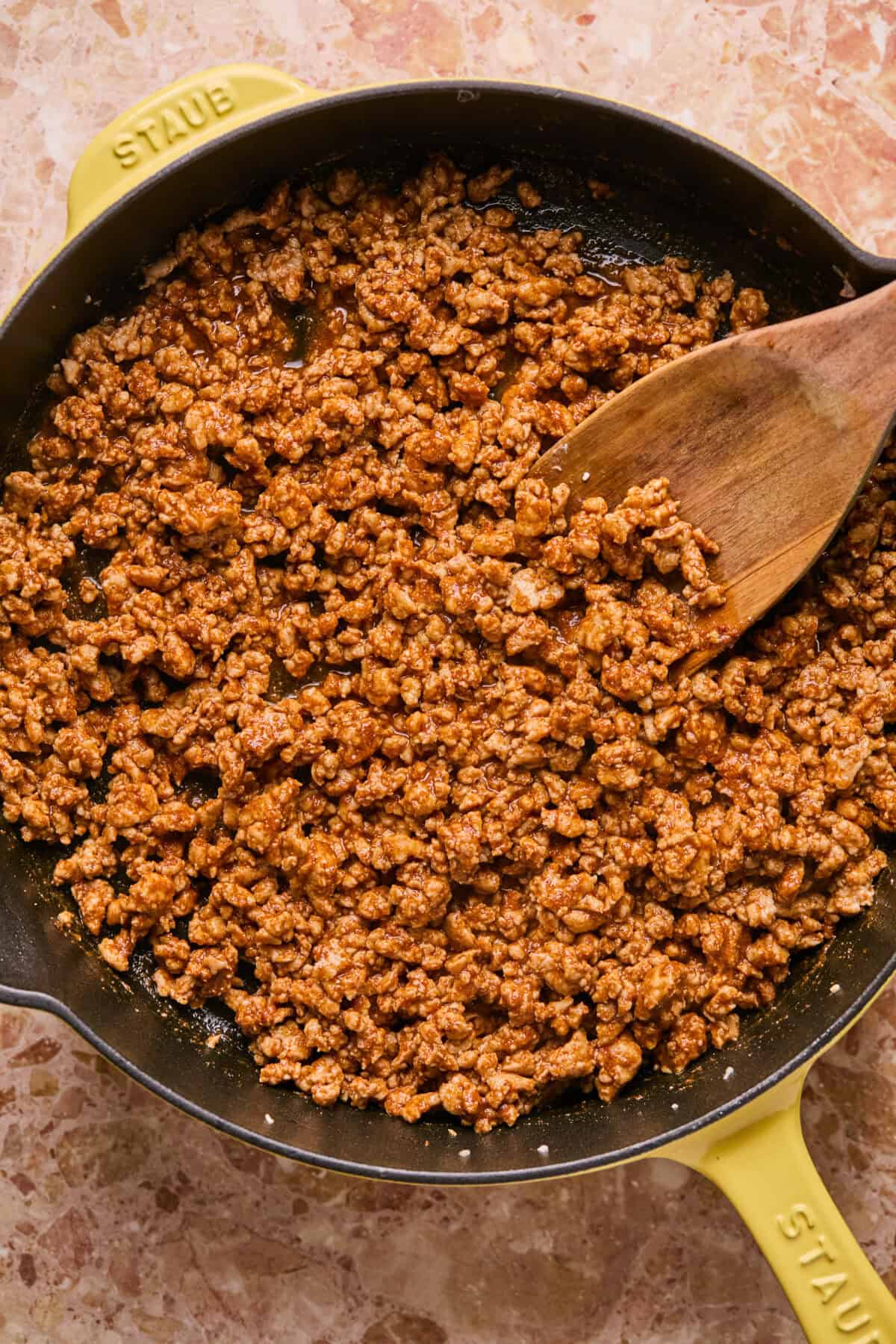 A pan filled with browned ground meat being stirred with a wooden spoon, reminiscent of the savory filling for baked chicken tacos. The yellow pan rests on a marbled countertop, and the meat appears seasoned and cooked, boasting a slightly oily texture.