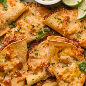 Close-up of crispy, golden-brown baked chicken tacos filled with melted cheese and topped with chopped cilantro. A small bowl of creamy green sauce and a few lime wedges are on the side. The tacos are arranged on a light green rectangular plate.