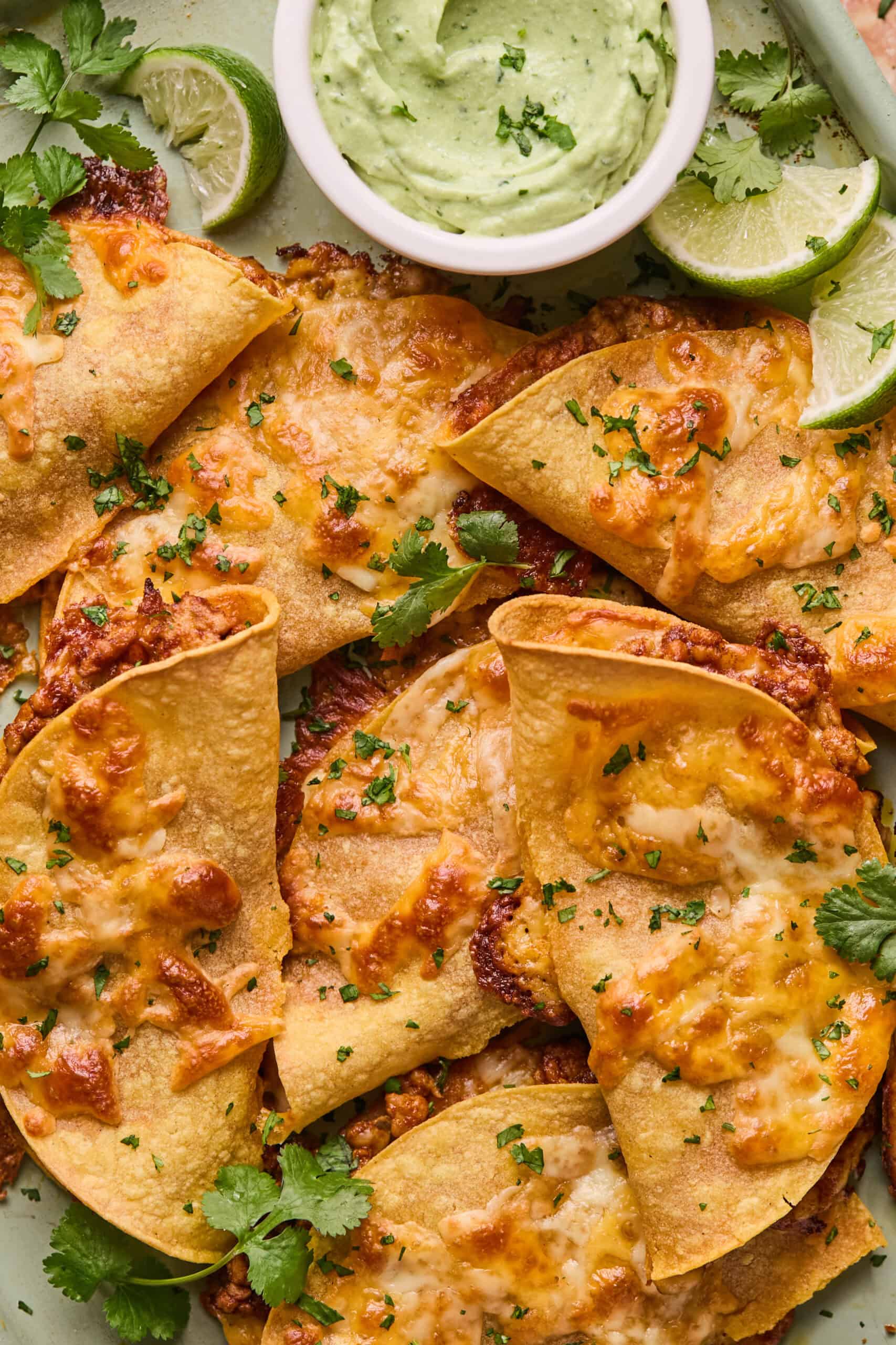 Close-up of crispy, golden-brown baked chicken tacos filled with melted cheese and topped with chopped cilantro. A small bowl of creamy green sauce and a few lime wedges are on the side. The tacos are arranged on a light green rectangular plate.