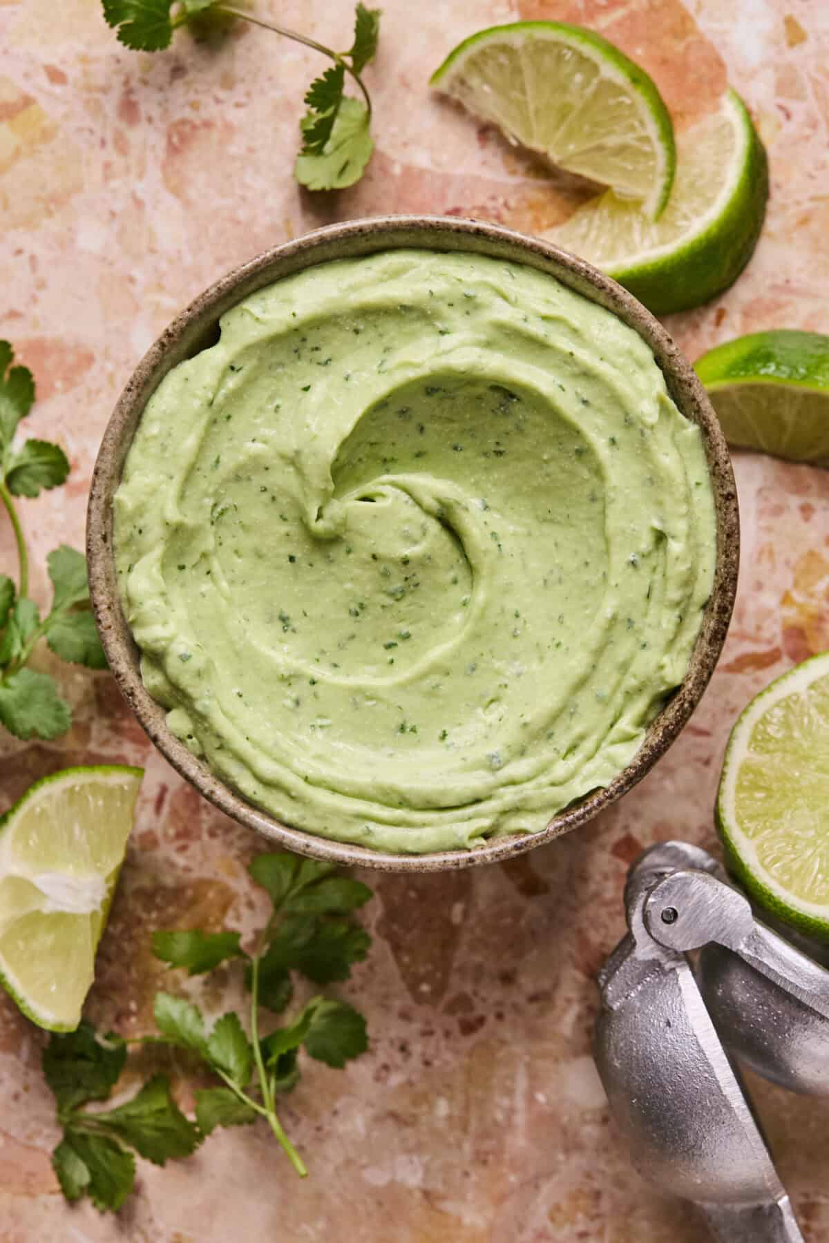 A bowl of creamy avocado crema garnished with cilantro sits on a marble surface. Nearby, lime wedges and a metal citrus squeezer add a zesty touch.