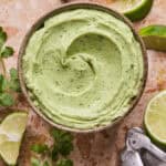 A bowl of creamy avocado crema garnished with cilantro sits on a marble surface. Nearby, lime wedges and a metal citrus squeezer add a zesty touch.