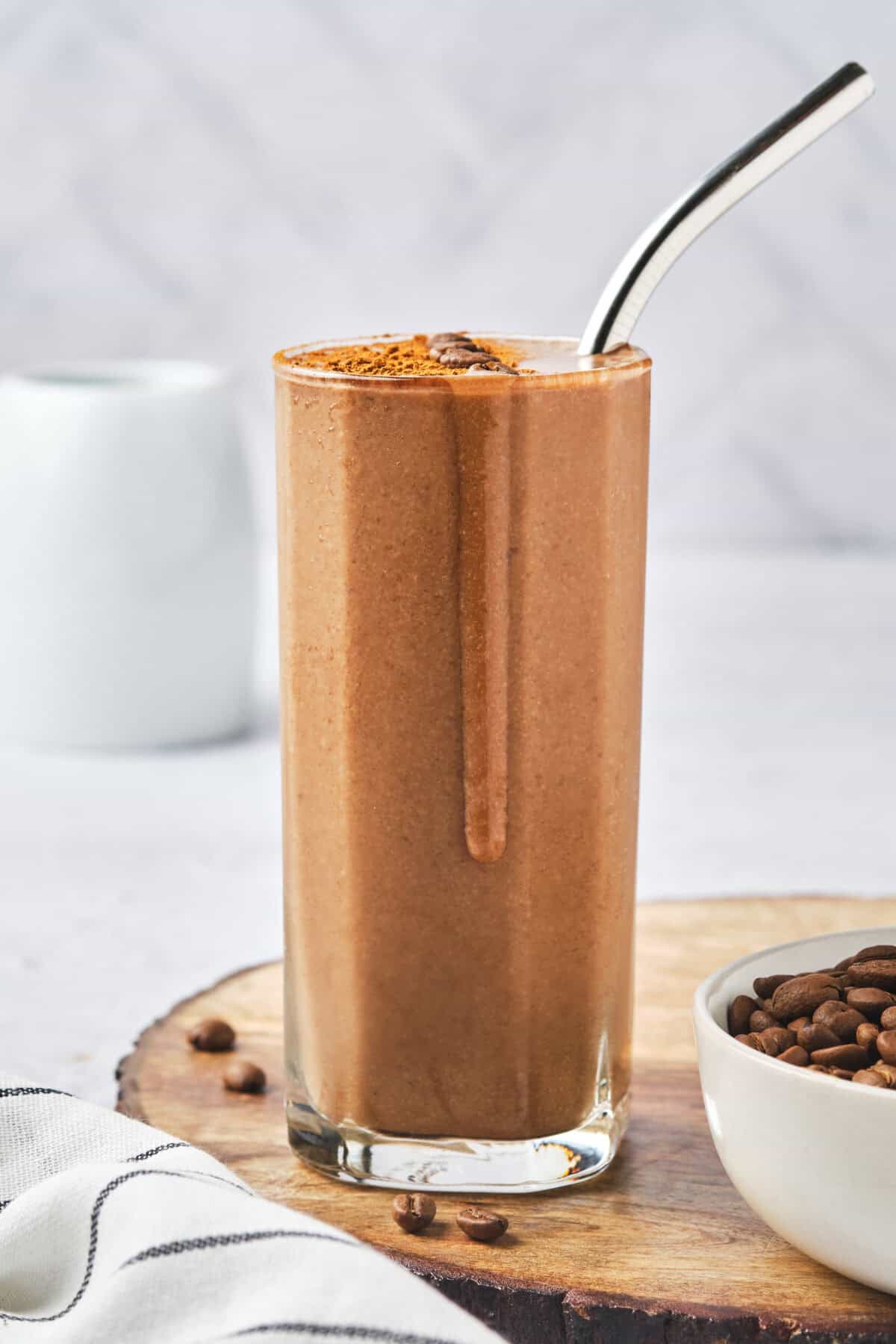 A tall glass of chocolate smoothie with a metal straw sits on a wooden surface, reminiscent of a rich coffee protein shake. The smoothie is topped with cocoa powder, and next to it is a small bowl filled with coffee beans, while a white mug is blurred in the background.