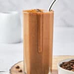 A tall glass of chocolate smoothie with a metal straw sits on a wooden surface, reminiscent of a rich coffee protein shake. The smoothie is topped with cocoa powder, and next to it is a small bowl filled with coffee beans, while a white mug is blurred in the background.