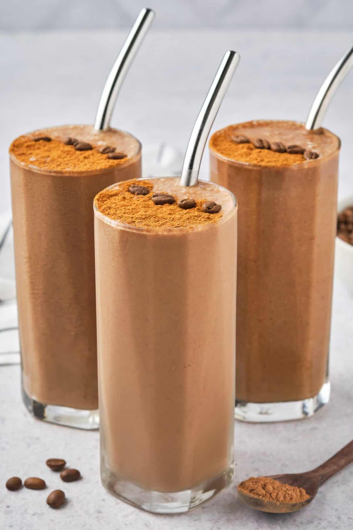 Three tall glasses brimming with creamy coffee protein smoothies, topped with cocoa powder and whole coffee beans. Each glass features a sleek metal straw. A wooden spoon dusted with cocoa powder sits in the foreground, while scattered coffee beans are artfully arranged on the table.