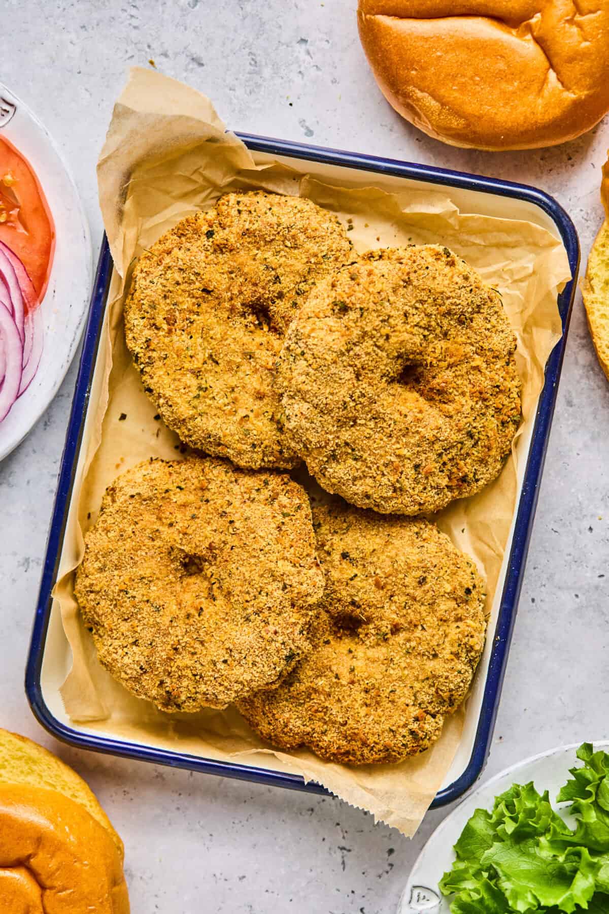 Four crispy air fryer chicken patties are arranged on a parchment-lined tray. The table also features sliced buns, leafy lettuce, and sliced red onions, suggesting burger preparations.