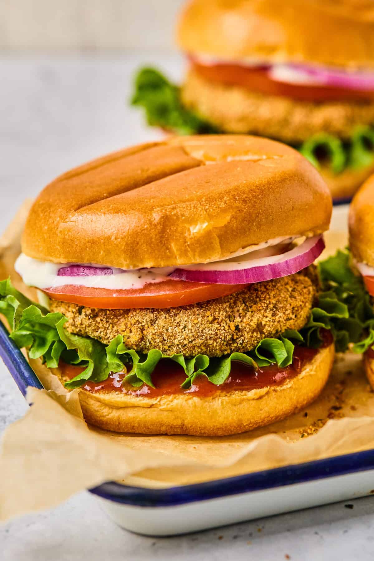 A close-up of a crispy fried green tomato sandwich on a bun with lettuce, tomato, red onion slices, and a dollop of creamy sauce. The sandwich is placed on parchment paper in a tray alongside air fryer chicken patties. Another sandwich is slightly visible in the background.