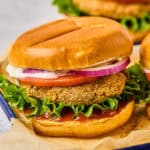 A close-up of a crispy fried green tomato sandwich on a bun with lettuce, tomato, red onion slices, and a dollop of creamy sauce. The sandwich is placed on parchment paper in a tray alongside air fryer chicken patties. Another sandwich is slightly visible in the background.