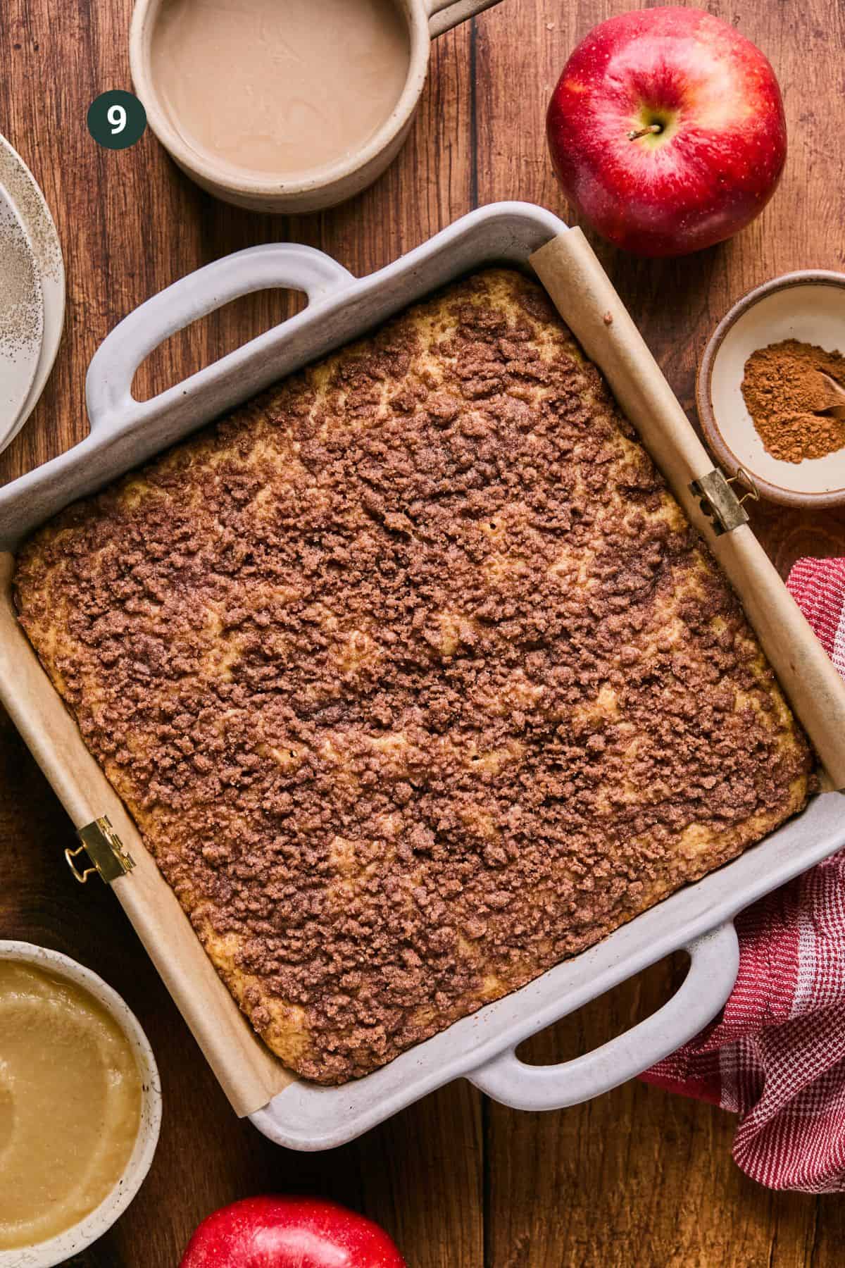 A freshly baked applesauce coffee cake sits in a square baking dish on a wooden table. Surrounding it are two red apples, a bowl of cinnamon, and a steaming cup of coffee, all nestled beside a charming red and white cloth.