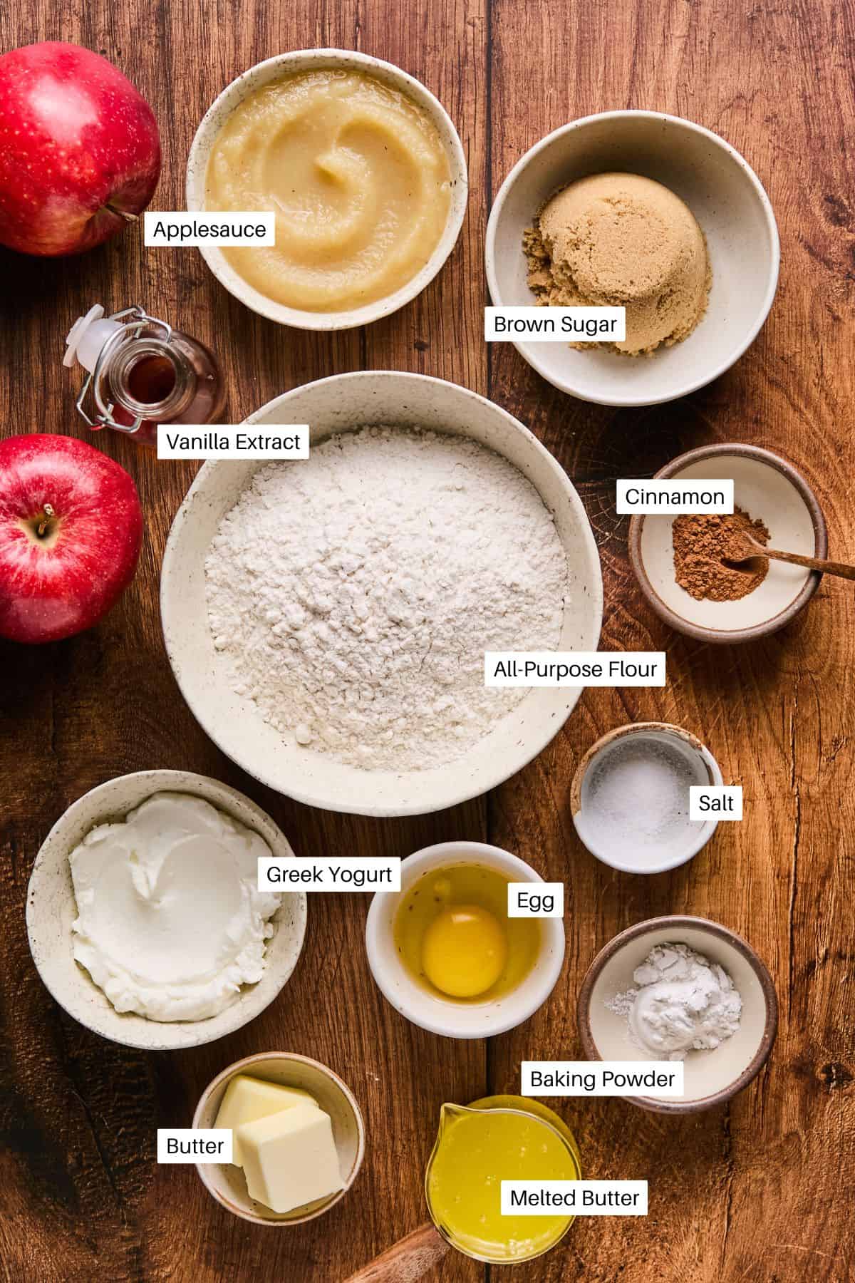 A wooden table showcases the essentials for whipping up a delicious applesauce coffee cake: bowls of brown sugar, vanilla extract, all-purpose flour, cinnamon, salt, Greek yogurt, an egg, baking powder, butter and melted butter. Two red apples sit beside them, ready to join the mix.