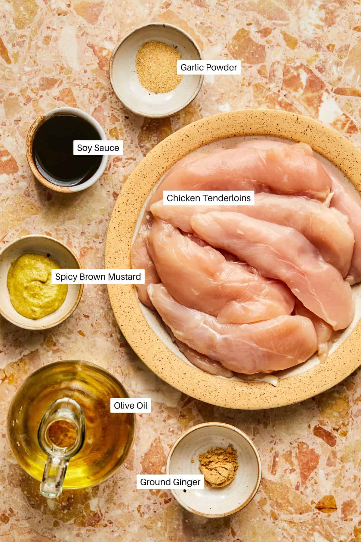 A ceramic bowl filled with raw chicken tenderloins is surrounded by small dishes containing soy sauce, spicy brown mustard, garlic powder, olive oil, and ground ginger on a marbled surface—perfect ingredients to prepare baked chicken tenderloins in the oven.