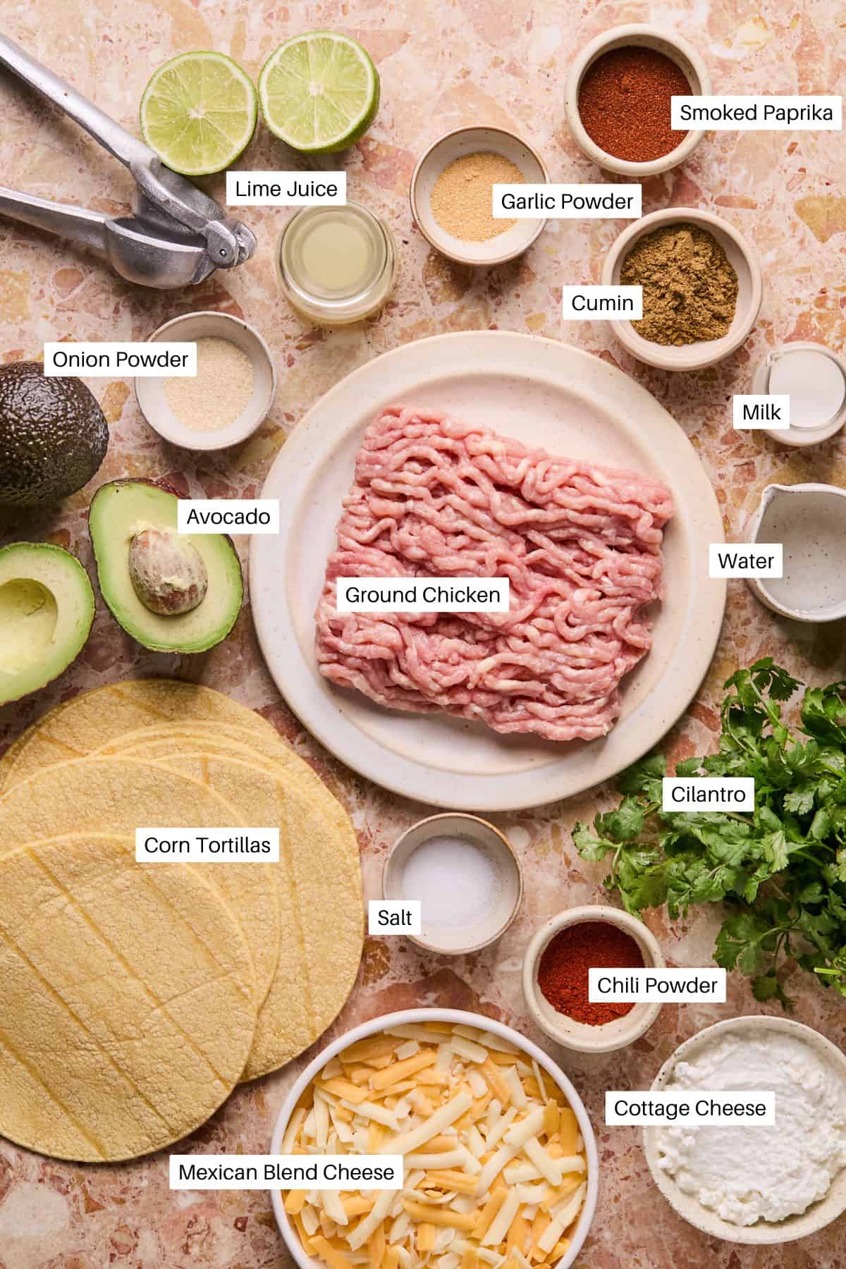 A flat lay of ingredients for baked chicken tacos, featuring ground chicken, avocado, corn tortillas, lime juice, various spices, cilantro, cheese, and cottage cheese. Each ingredient is conveniently labeled for easy preparation.