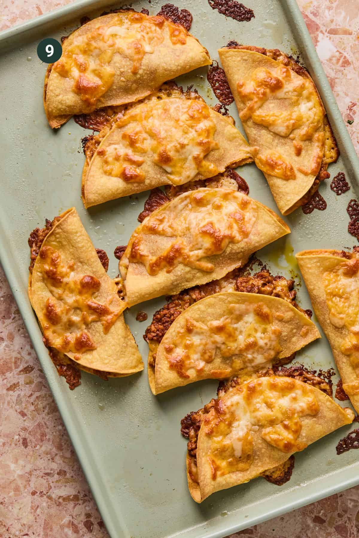 A baking sheet with nine crispy baked chicken tacos filled with a cheesy, browned mixture. The tacos are golden and slightly charred, resting on a light green tray against a marbled surface.