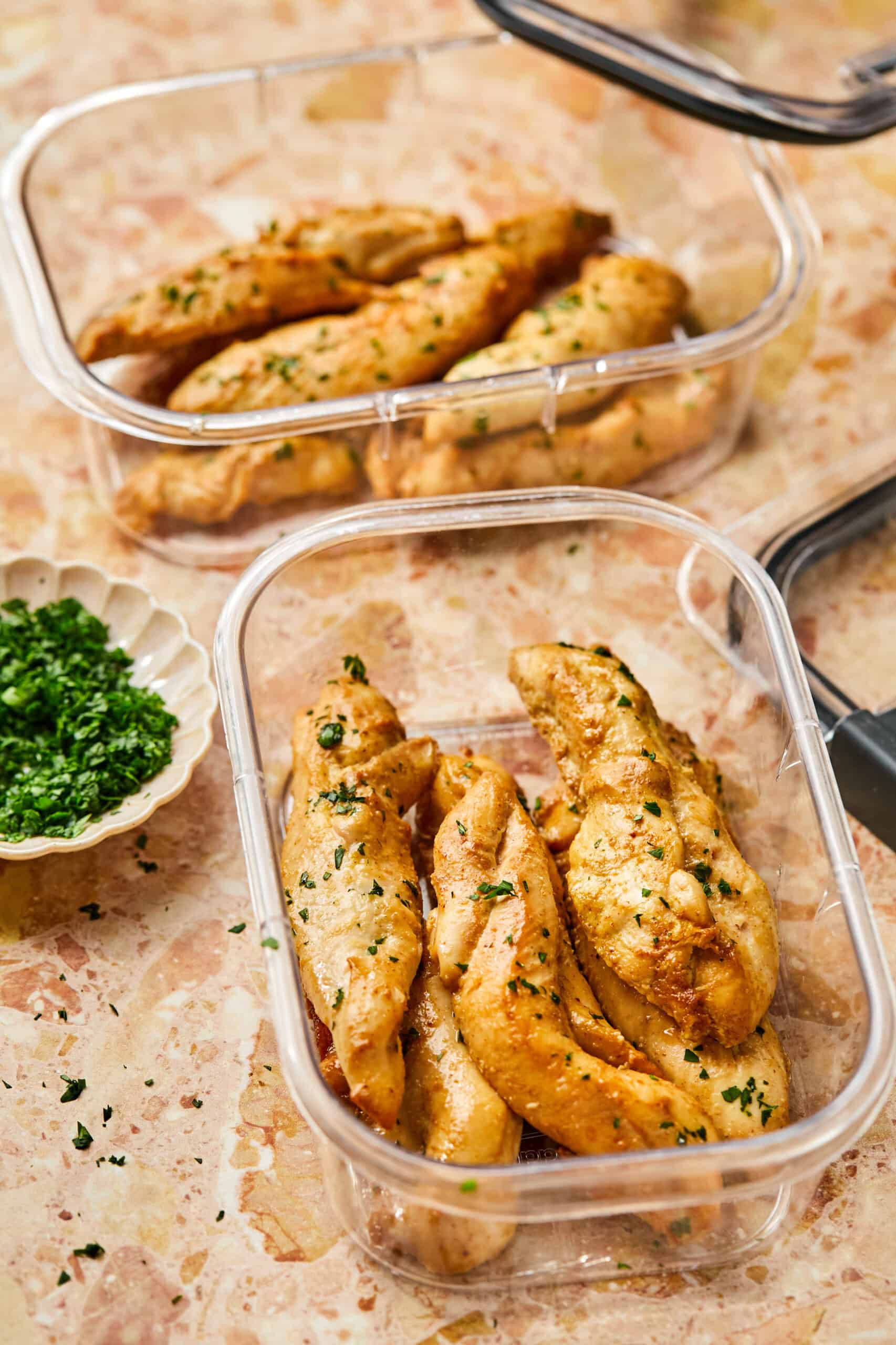 Two clear containers filled with baked chicken tenderloins are placed on a marble surface. A small dish of chopped parsley is beside them. The oven-cooked chicken, seasoned and garnished with herbs, offers a fresh and appetizing look.