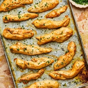 Golden-brown baked chicken tenderloins in the oven are sprinkled with fresh herbs and arranged on a baking sheet. A small bowl of chopped green herbs sits nearby, adding a touch of color to the marble surface beneath.