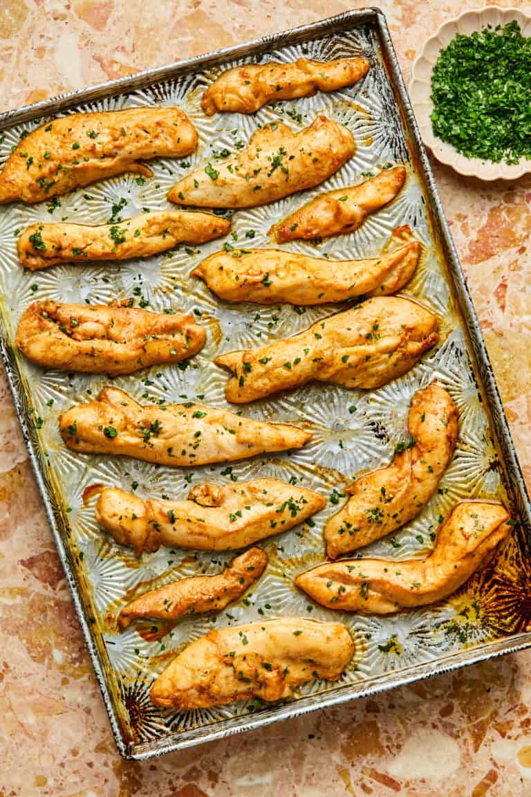 Golden-brown baked chicken tenderloins in the oven are sprinkled with fresh herbs and arranged on a baking sheet. A small bowl of chopped green herbs sits nearby, adding a touch of color to the marble surface beneath.