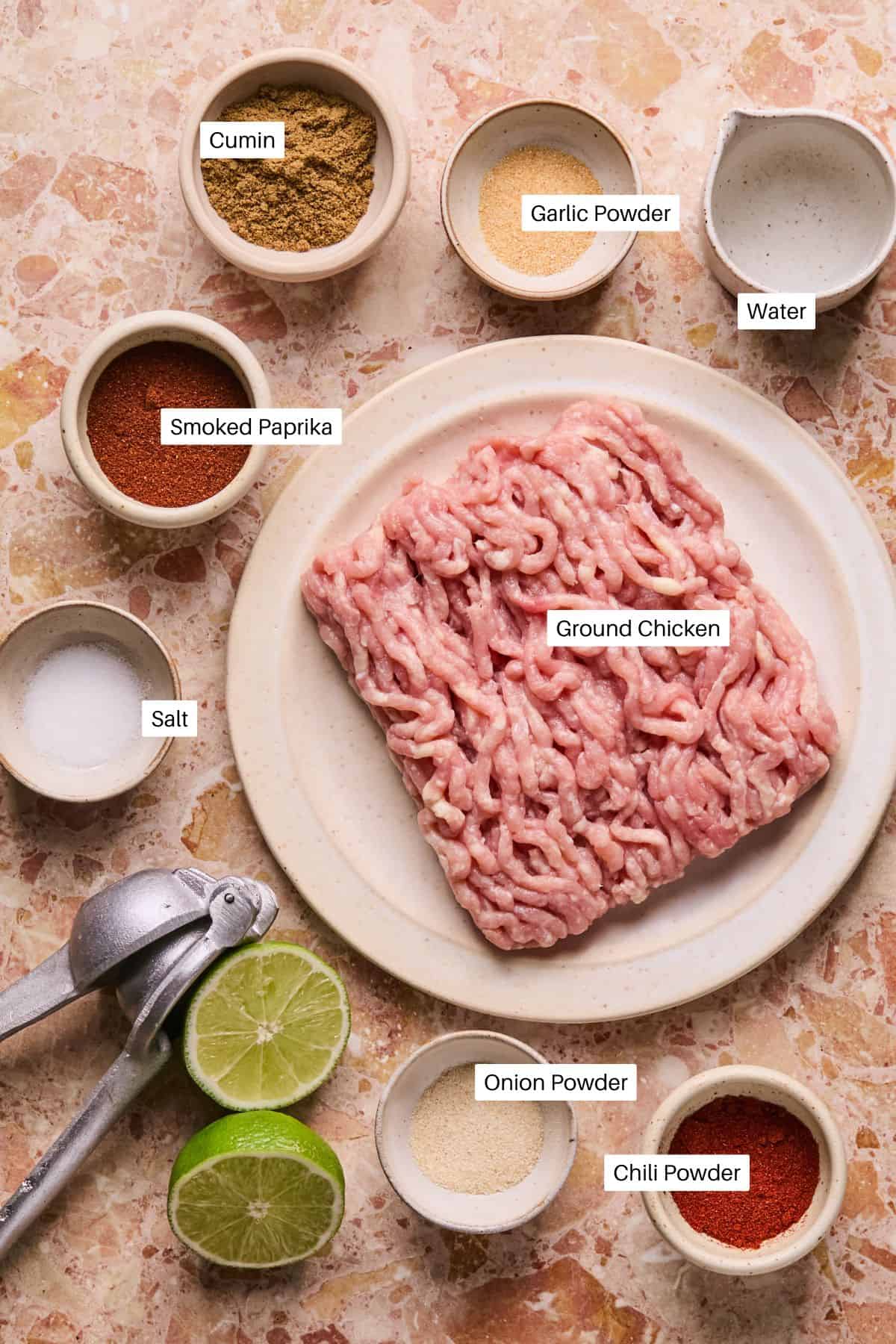 A plate of ground chicken, destined for baked chicken tacos, sits amidst bowls of cumin, garlic powder, smoked paprika, salt, onion powder, chili powder, and water. A garlic press and halved limes rest on the textured surface nearby.