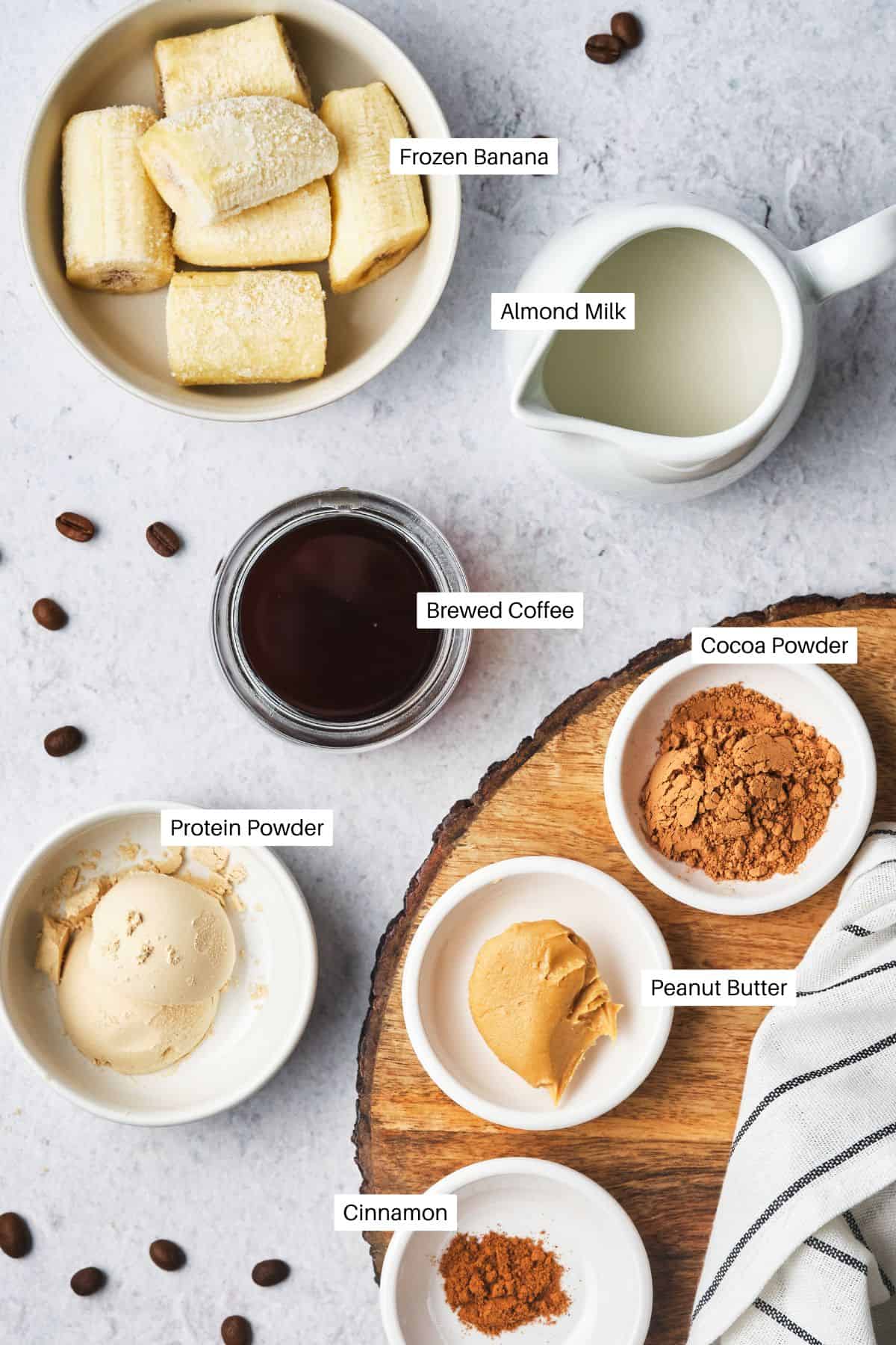 An image of coffee protein shake ingredients: a bowl of frozen banana slices, a jug of almond milk, brewed coffee in a jar, small bowls with cocoa powder, protein powder, peanut butter, and a sprinkle of cinnamon. Coffee beans are scattered on the surface.