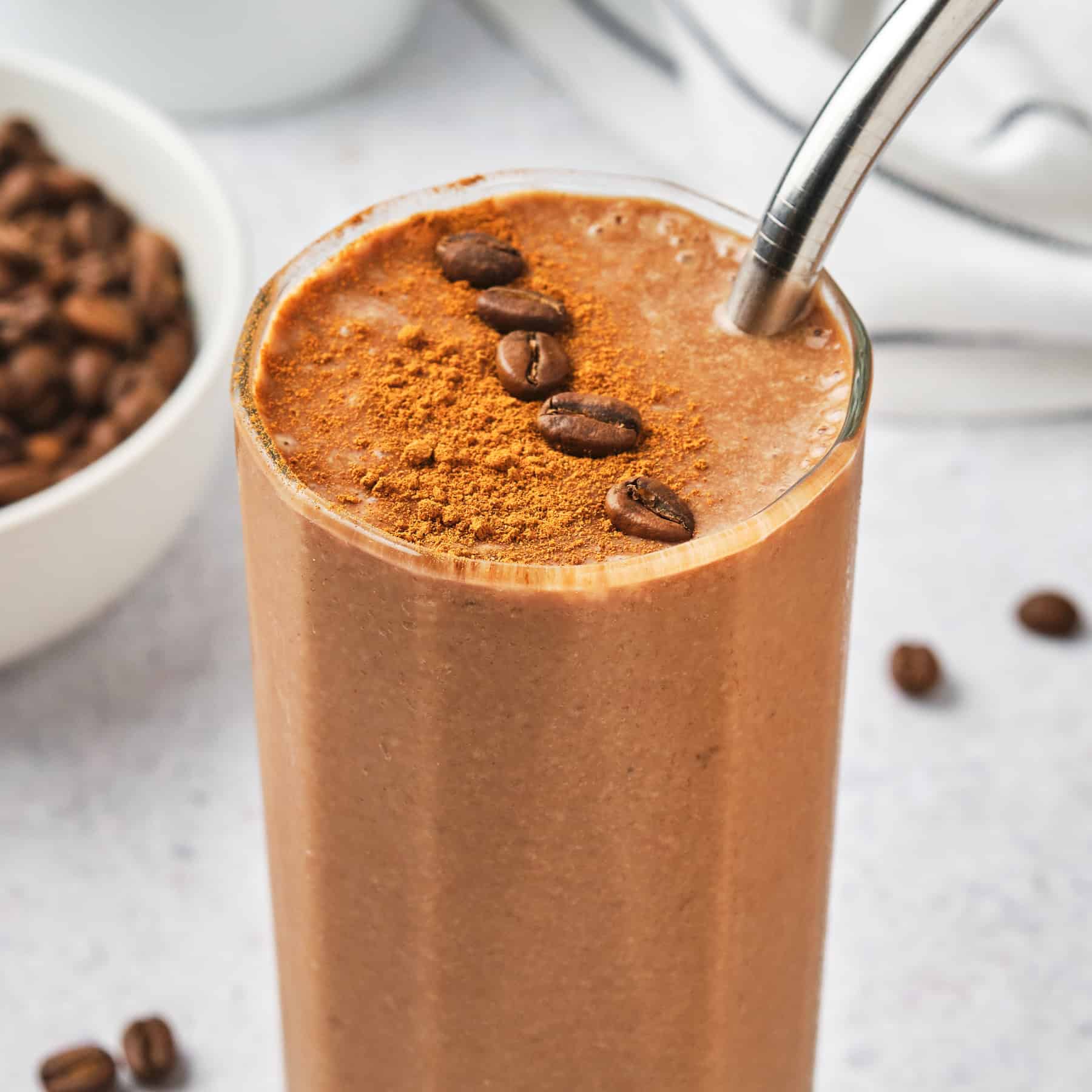 A tall glass brimming with a creamy Coffee Protein Shake is topped with cocoa powder and coffee beans. A metal straw invites you to sip this energizing delight. In the background, there's a bowl of coffee beans and a striped cloth, adding to the cozy ambiance.