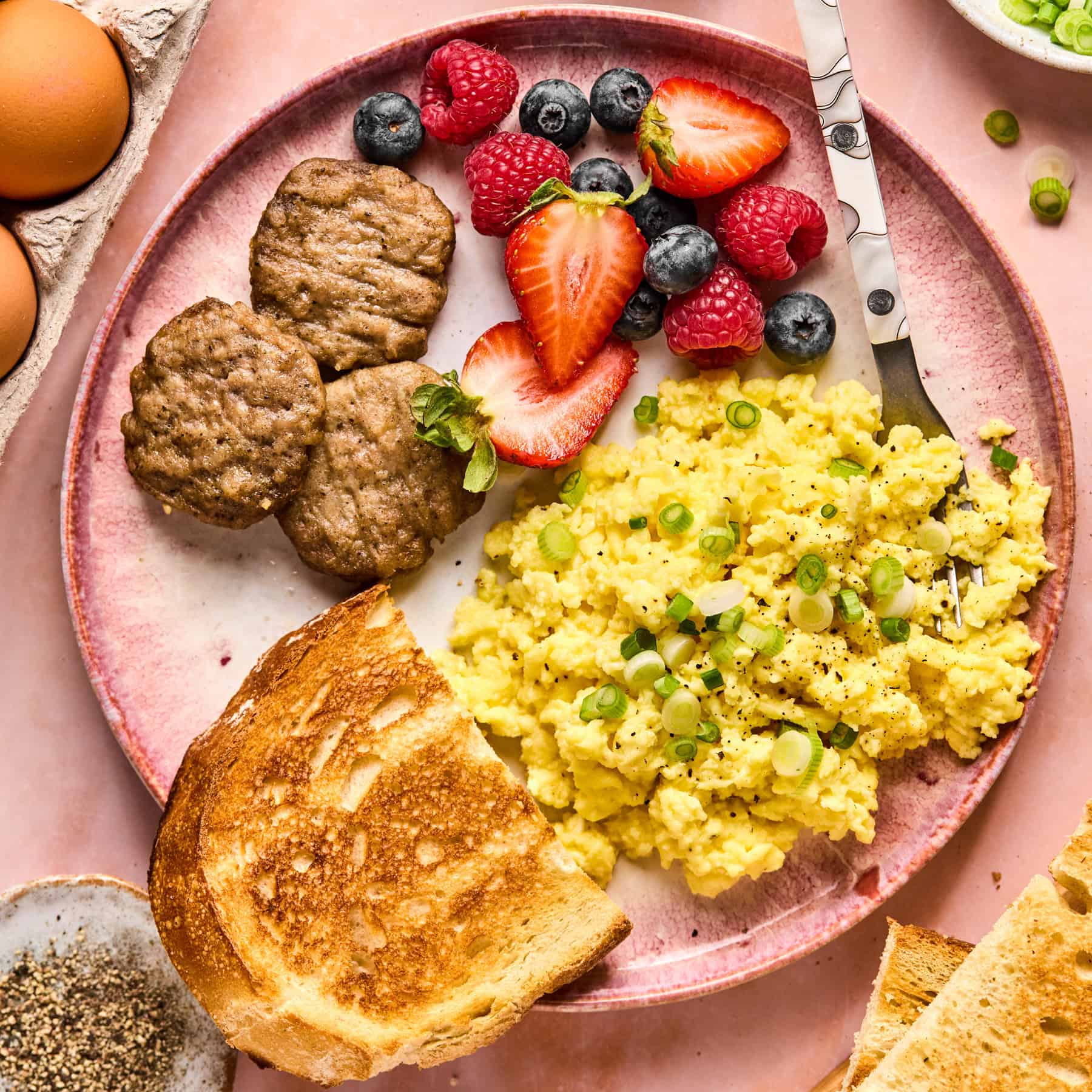 A breakfast plate with scrambled eggs and cottage cheese garnished with green onions, sausage patties, fresh strawberries, blueberries, raspberries, and a slice of toasted bread. Eggs in shells and a bowl of pepper are beside the pink plate.