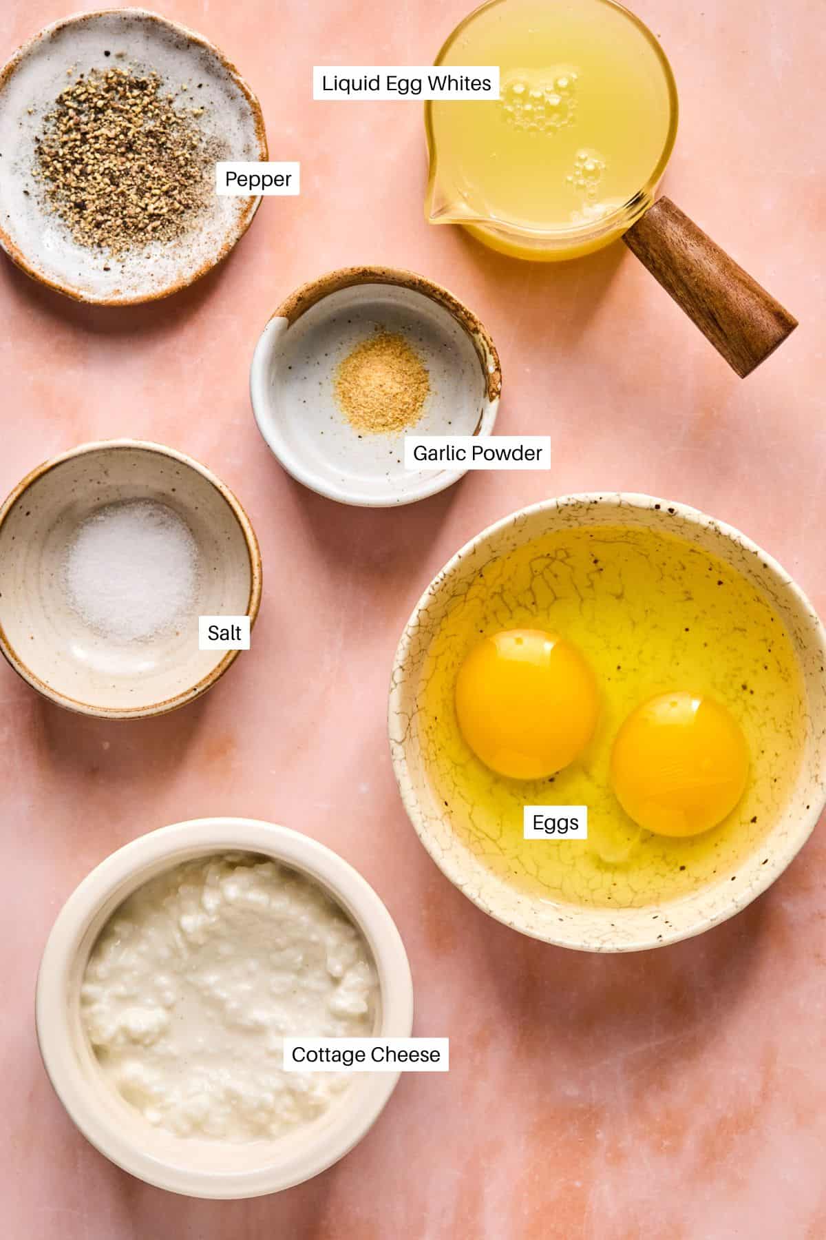 An assortment of ingredients featuring cottage cheese and eggs: liquid egg whites in a jug, two eggs in a bowl, small bowls of pepper, garlic powder, and salt, all elegantly placed on a pink surface.