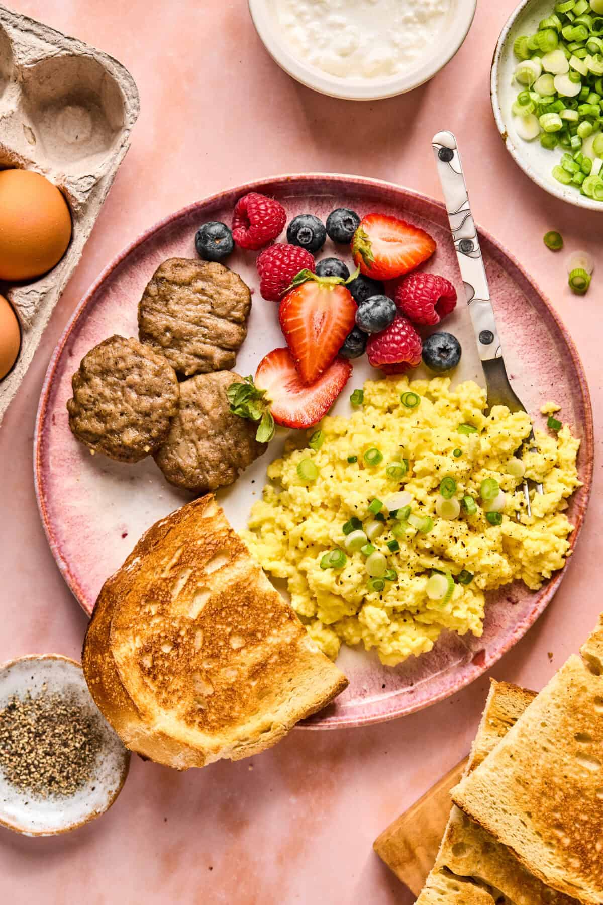 A breakfast plate featuring scrambled eggs garnished with green onions, alongside sausage patties, crispy toast, and mixed berries like strawberries, raspberries, and blueberries. Nearby are whole eggs, a small bowl of seasoning, sliced green onions, and a dollop of creamy cottage cheese.