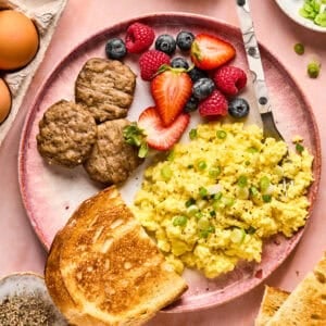 A breakfast plate featuring scrambled eggs garnished with green onions, alongside sausage patties, crispy toast, and mixed berries like strawberries, raspberries, and blueberries. Nearby are whole eggs, a small bowl of seasoning, sliced green onions, and a dollop of creamy cottage cheese.