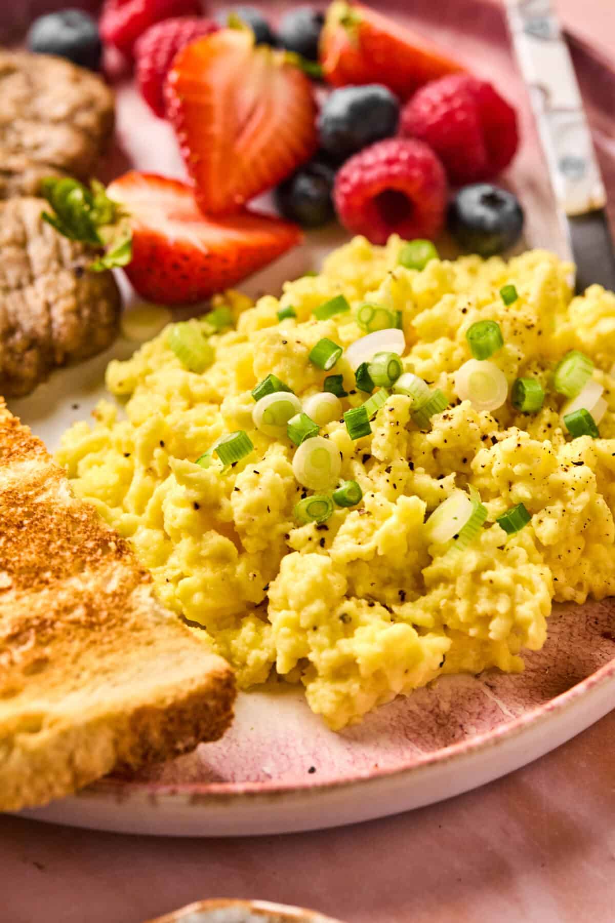 A plate of breakfast food featuring scrambled eggs topped with green onions, a slice of toast, and a medley of strawberries, raspberries, and blueberries. A steak patty joins the ensemble on the pinkish surface. For added delight, a dollop of cottage cheese accompanies the eggs.