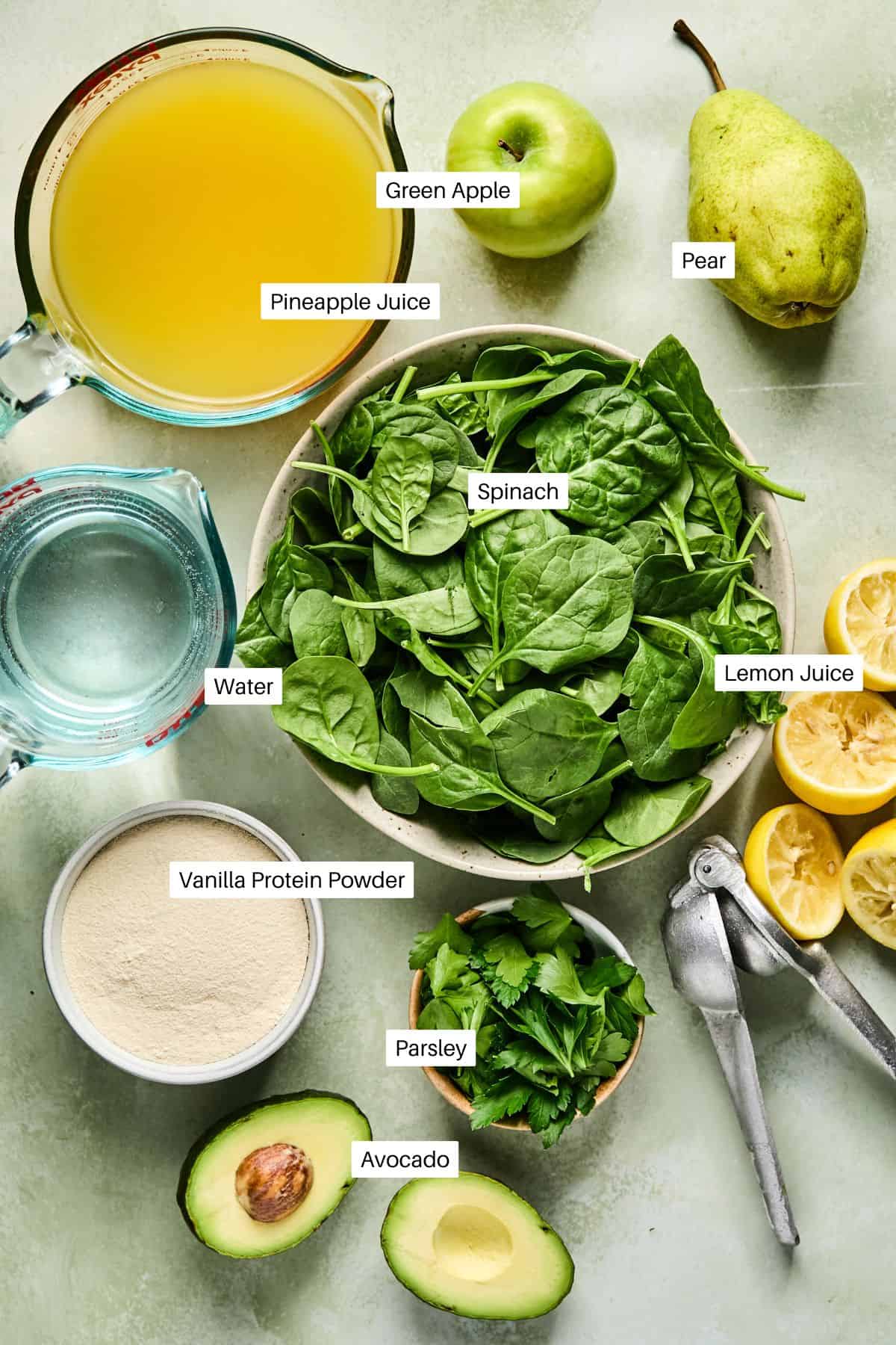 Flat lay of Green Protein Smoothie ingredients labeled on a light surface. Includes spinach, lemon juice, pear, green apple, pineapple juice, water, vanilla protein powder, parsley, and avocado. A glass jar and wooden board are partially visible.