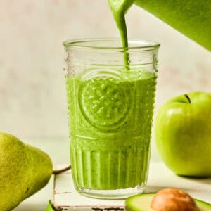 A clear glass is being filled with a green protein smoothie from a pitcher. Surrounding the glass are a whole pear, a green apple, spinach leaves, and a halved avocado, all on a light background.