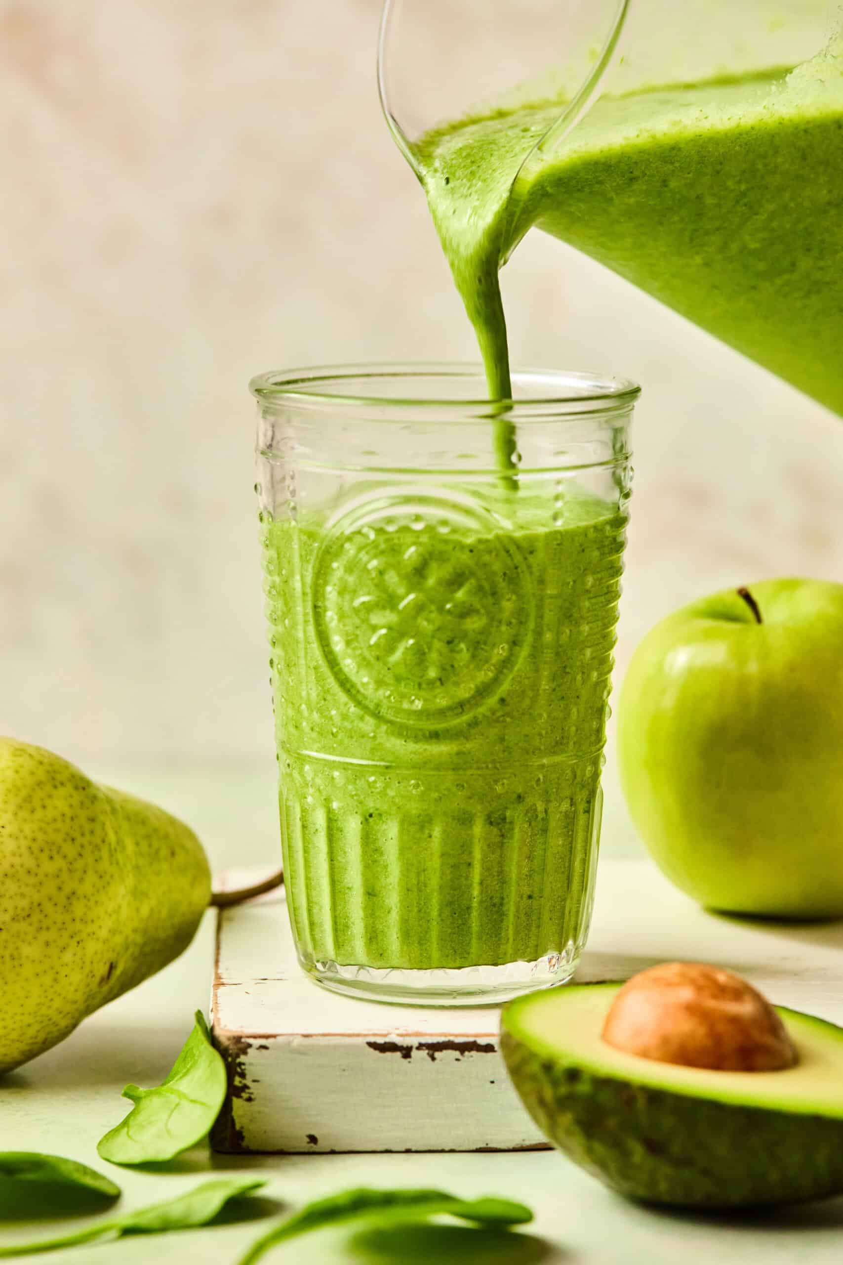 A clear glass is being filled with a green protein smoothie from a pitcher. Surrounding the glass are a whole pear, a green apple, spinach leaves, and a halved avocado, all on a light background.