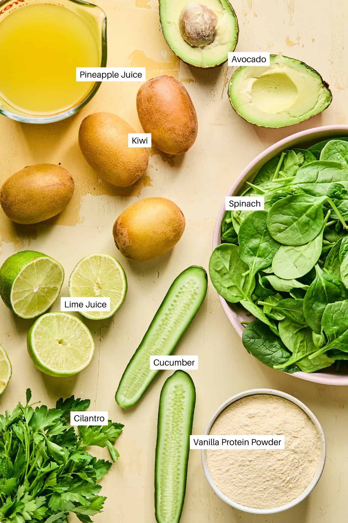 An assortment of smoothie bowl ingredients on a light surface, featuring pineapple juice, whole kiwi, halved avocado, spinach in a bowl, lime juice, cucumber slices, cilantro, and vanilla protein powder. Each item is labeled for a perfect blend.