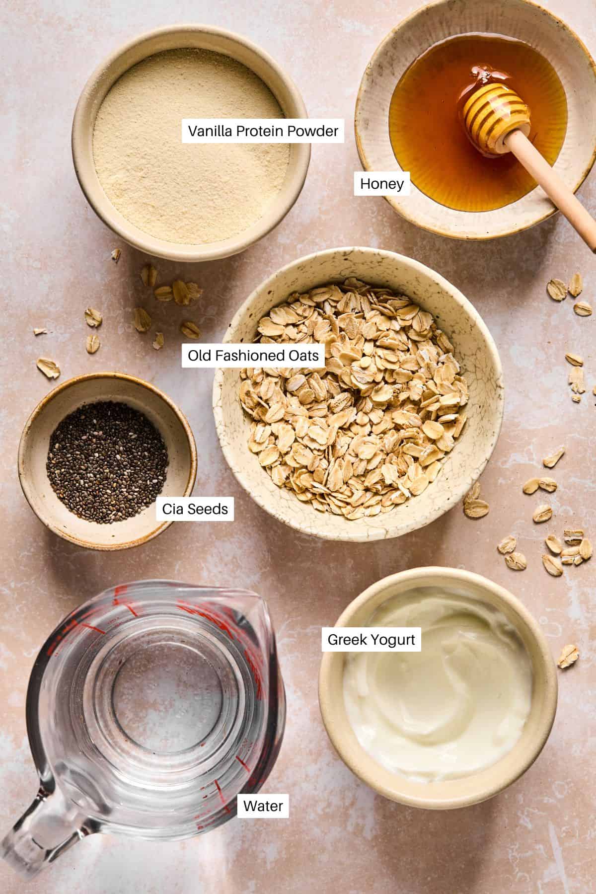 Overhead view of ingredients for an overnight oats recipe: vanilla protein powder, honey, old-fashioned oats, chia seeds, Greek yogurt, and water are neatly arranged in separate bowls or containers on a light surface.