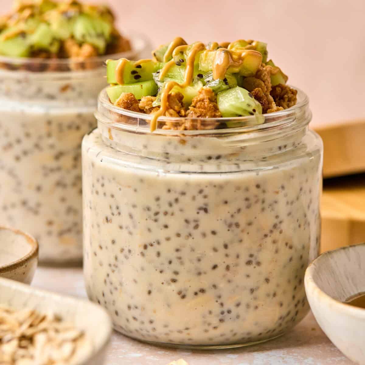 Two jars filled with chia pudding topped with chopped kiwi, granola, and a drizzle of peanut butter evoke a healthy breakfast vibe. Set against a soft pink background with oats scattered around, these jars pair perfectly with overnight oats made with water for an energizing start to the day.