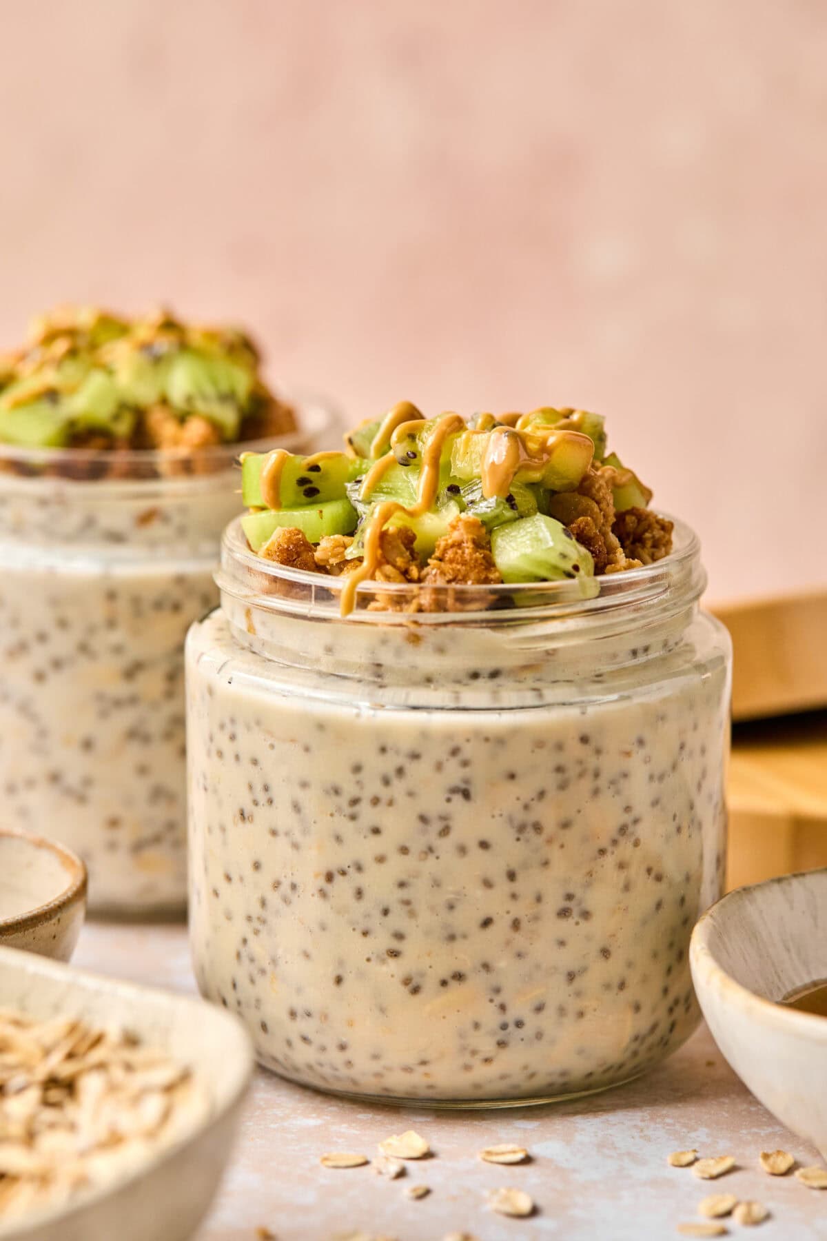 Two jars of chia pudding topped with granola, green fruit pieces, and a drizzle of nut butter. Next to the jars, you'll find bowls filled with overnight oats made with water. Everything is set on a light surface, while the background remains softly blurred.