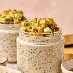 Two jars of chia pudding topped with granola, green fruit pieces, and a drizzle of nut butter. Next to the jars, you'll find bowls filled with overnight oats made with water. Everything is set on a light surface, while the background remains softly blurred.