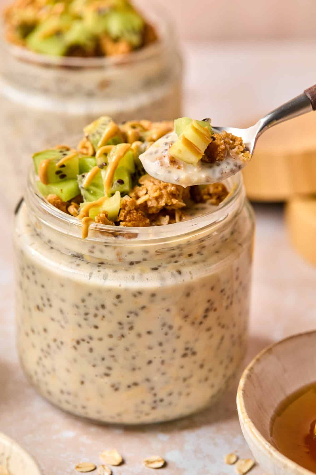 A jar of creamy chia pudding topped with sliced kiwi, granola, and a drizzle of nut butter sits beside another filled with overnight oats made with water. A spoon is scooping out a portion, while another jar and a bowl are partially visible in the background.