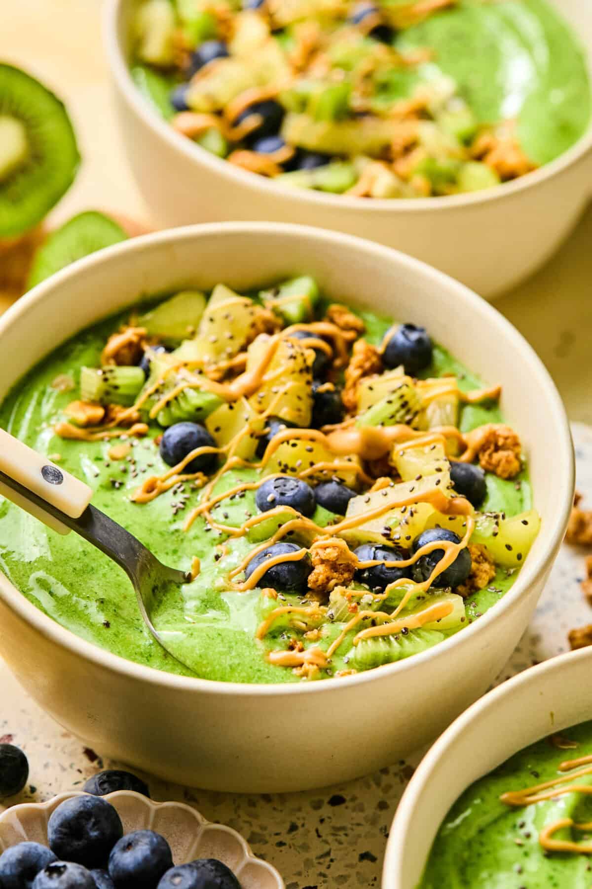 A vibrant smoothie bowl brimming with a green blend, crowned with juicy blueberries, kiwi slices, crunchy granola, and a drizzle of peanut butter. A spoon waits eagerly in the bowl, while more bowls and ingredients add charm to the background.