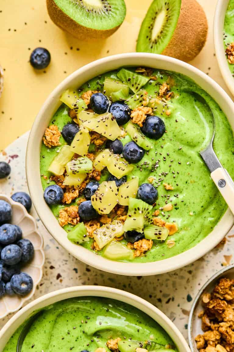 A vibrant green smoothie bowl topped with fresh kiwi chunks, blueberries, granola, and chia seeds. Surrounding the smoothie bowl are whole kiwi fruits and more blueberries on a speckled surface. A spoon rests invitingly in the bowl.