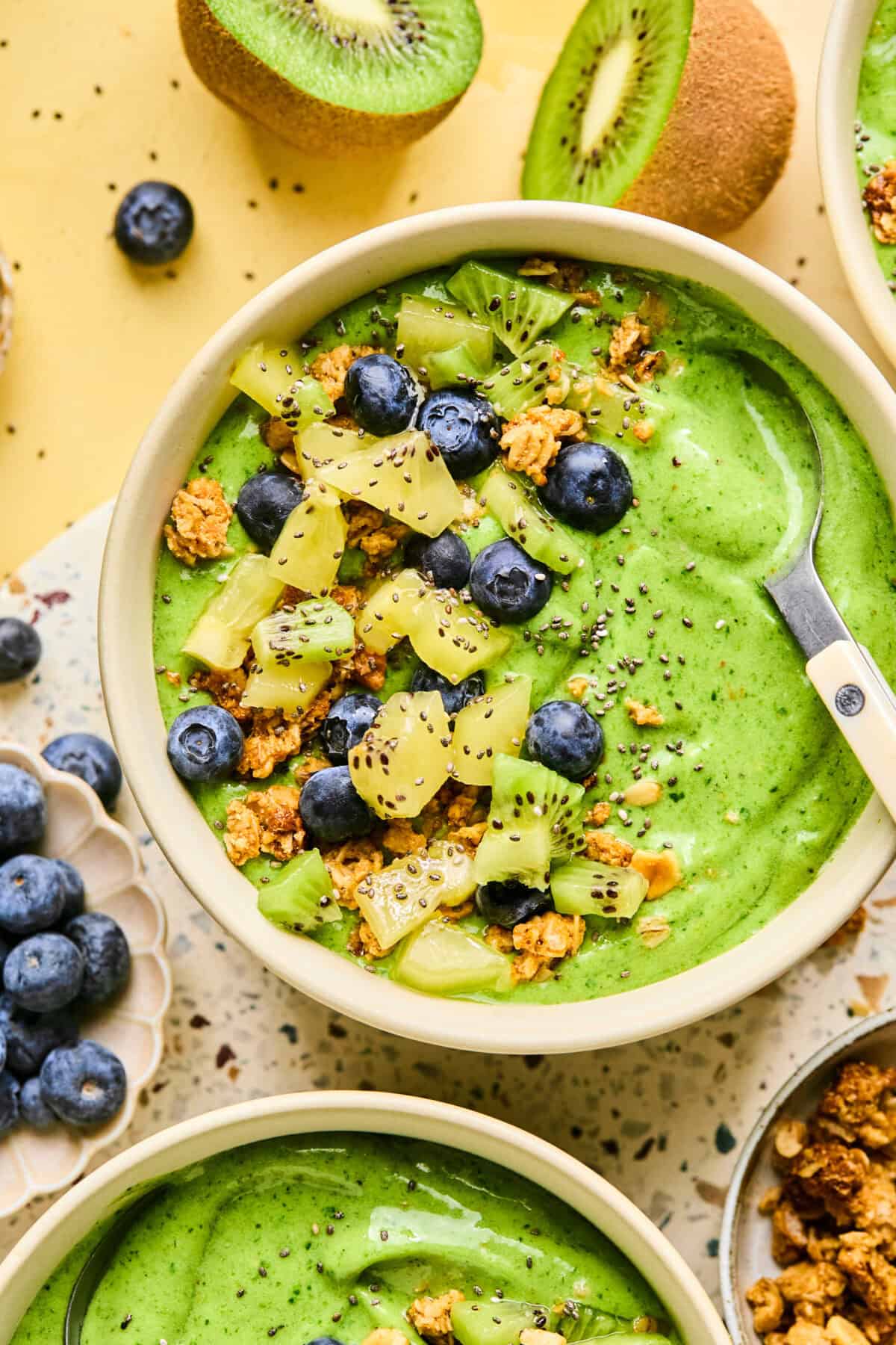 A vibrant smoothie bowl brims with lush green hues, adorned with sliced kiwi, blueberries, granola, and chia seeds. A spoon rests on the bowls edge. Whole kiwis and a small bowl of blueberries complete the scene on a speckled surface.